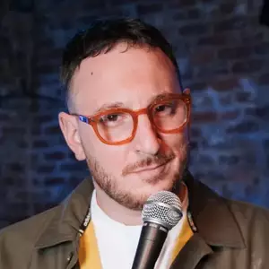 Comedian Ahri Findling poses with a microphone in front of a brick wall