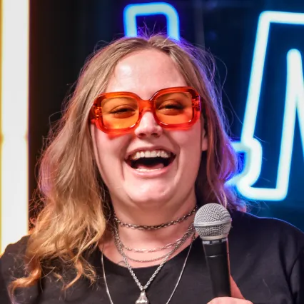 Liz_Splatt Comedian Liz Splatt poses on stage in front of the LMAOF logo.
