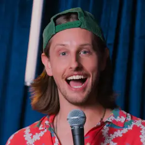 Comedian Austin Kress smiles to camera in front of a blue curtain.