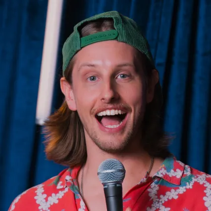 Comedian Austin Kress smiles to camera in front of a blue curtain.