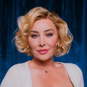 Comedian Billie Lee smirks to camera in front of a blue curtain.