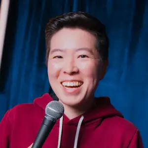Comedian Irene Tu smiles to camera in front of a blue curtain.