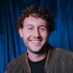 Comedian JT Tomlinson smiles to camera in front of a blue curtain.