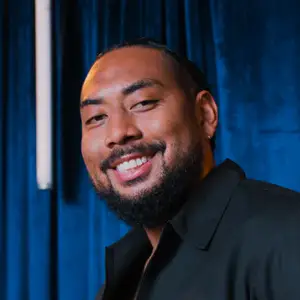 Comedian Keith Johnson smiles to camera in front of a blue curtain.
