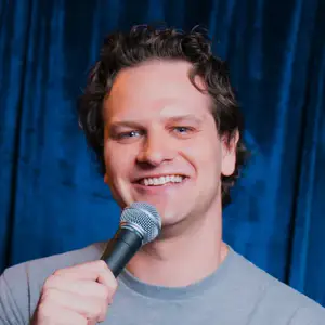 Comedian Kyle Rehl smiles to camera in front of a blue curtain.