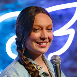 Comedian Maggie Lalley poses on stage in front of the OnlyFans logo, with mic, at LMAOF 110 in New York