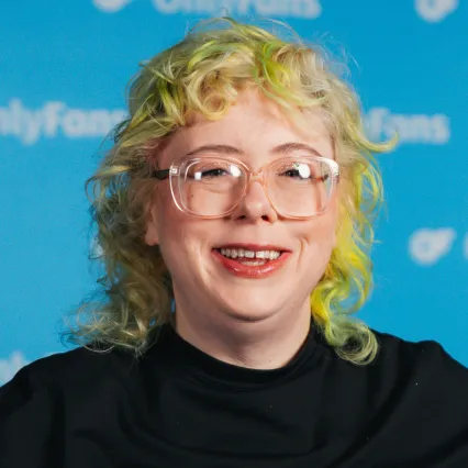 Comedian Alison Spittle smiles toward the camera in front of a blue OnlyFans backdrop.