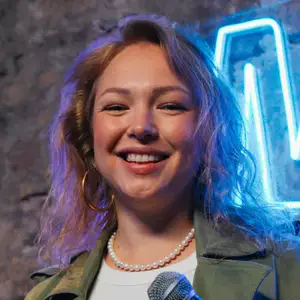 Kristina Pilska poses with a microphone in front of a neon OnlyFans sign on a brick wall.
