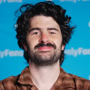 Paddy Young smirks to camera in front of a blue OnlyFans sponsored step and repeat.