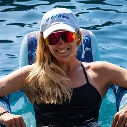 Steph Clutterbuck relaxing on a pool float wearing an OnlyFans cap and reflective sunglasses.