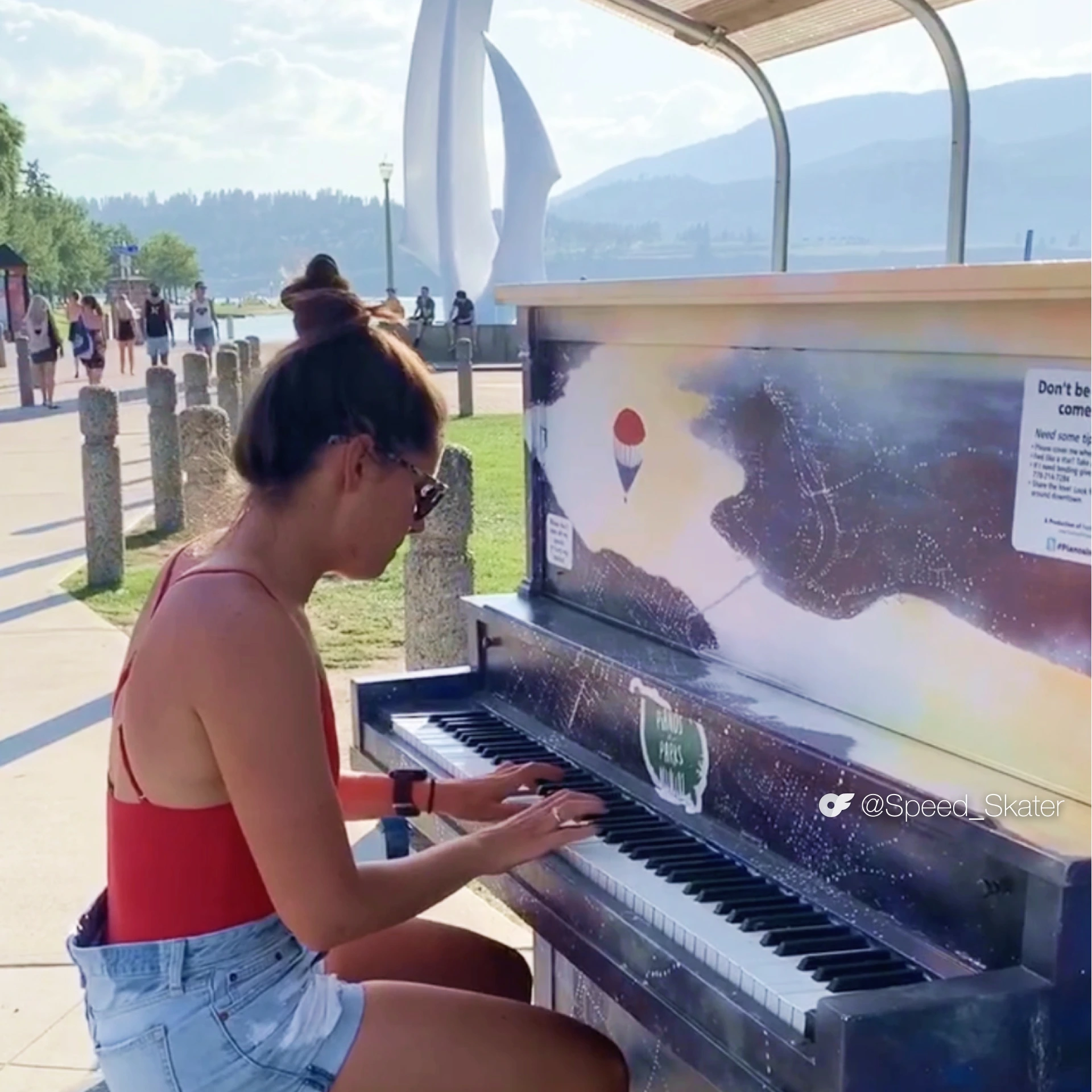 Alexandra Ianculescu plays a public outdoor piano near a waterfront, wearing a red tank top and denim shorts with mountains and trees in the background.