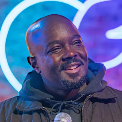 Reg Thomas on stage at a comedy club, reacting mid-joke with an expressive look.