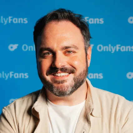 Sam Wiles, comedian, smiles in front of a blue backdrop with a repeating OnlyFans logo.