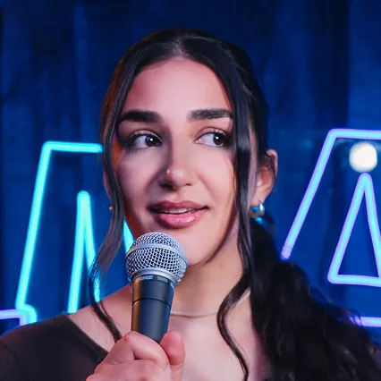 Char Ramtin, comedian, poses with a microphone in front of a neon sign.