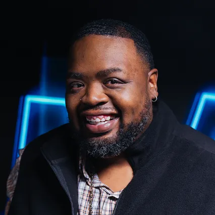 Charlie Mac, comedian, smiles in front of a neon sign.