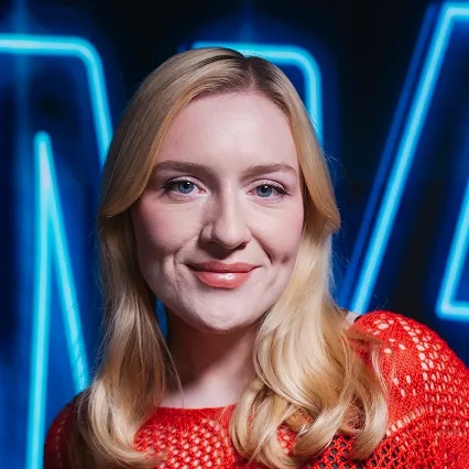 Hannah Jones, comedian, smirks in front of a neon sign.