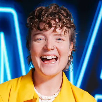 Rachel Scanlon, comedian, smiles in front of a neon sign.
