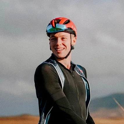 CallumStedman Triathlete Callum Stedman smiling outdoors in cycling gear, wearing a helmet and sunglasses perched on top, with a blurred natural landscape in the background.