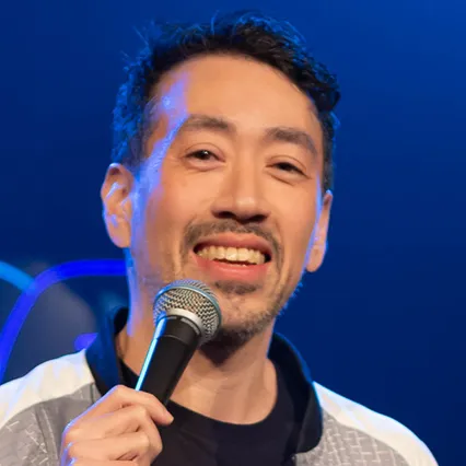 Stand-up comedian Jerry Tran smiling on stage while holding a microphone, lit with blue stage lighting and facing the audience.