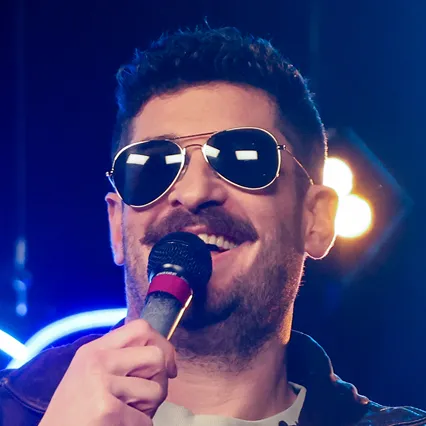 Stand-up comedian Timmy No Brakes holds a microphone and smiles as he performs on stage in aviator sunglasses.