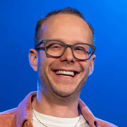 TylerFowler Stand-up comedian Tyler Fowler smiling on stage under blue lighting, wearing glasses and a casual jacket while facing the audience.