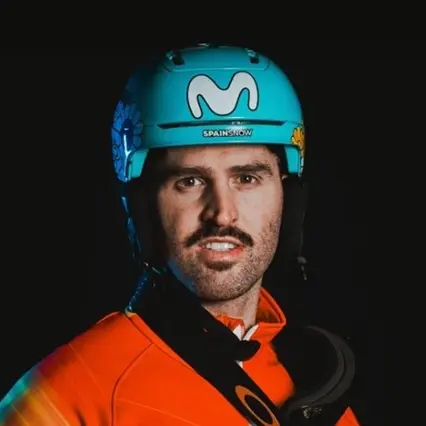 Lucas Eguibar, professional snowboard cross rider, wears an orange racing top and blue helmet while posing against a black backdrop.
