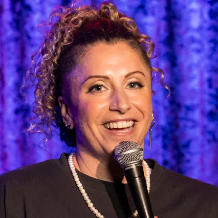 AdrienneFish Adrienne Fish, stand-up comedian, smiles while holding a microphone onstage.