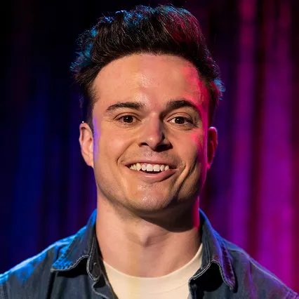 AndrewBarr Andrew Barr, stand-up comedian, smiles in a portrait against a purple and blue stage backdrop.