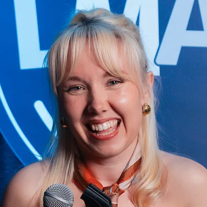 BeckyUmbers Becky Umbers, comedian, poses with a microphone in front of the LMAOF logo.