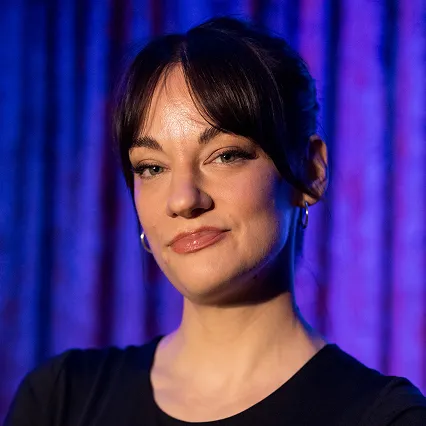 Hannah Veldhoen, stand-up comedian, poses for a portrait against a purple and blue stage backdrop.