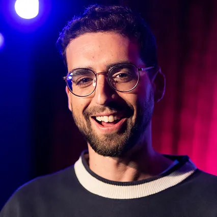 Jacob Balshin, stand-up comedian, smiles in a portrait against a purple and blue stage backdrop.