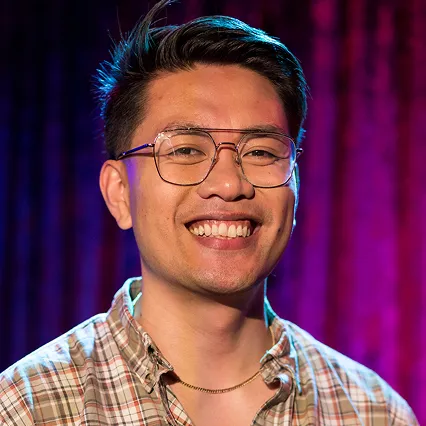 JamesRoque James Roque, stand-up comedian, smiles in a portrait against a purple and blue stage backdrop.
