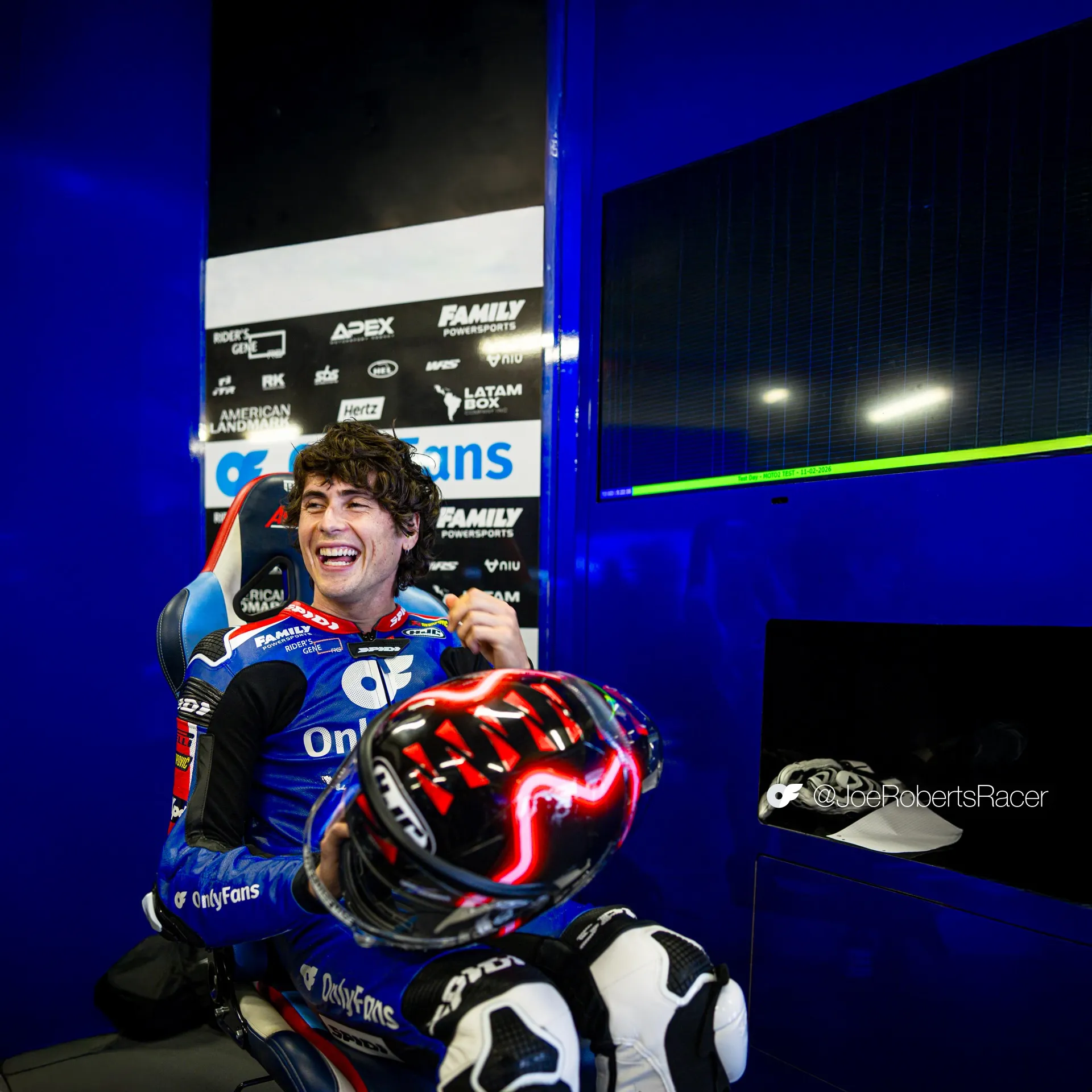 OART's Joe Roberts sits in the OnlyFans American Racing Team garage holding his helmet and smiling in full racing leathers.