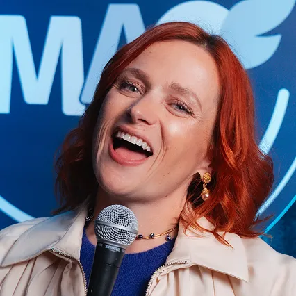 Kelly Ford, comedian, poses with a microphone in front of the LMAOF logo.