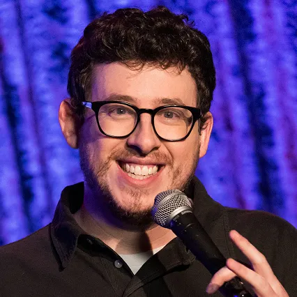 Mayce Galoni, stand-up comedian, smiles while holding a microphone onstage.