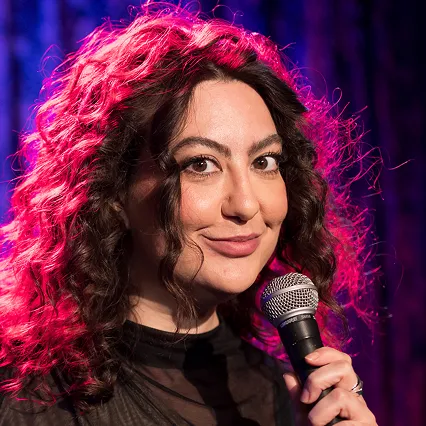 Natalie Norman, stand-up comedian, smiles while holding a microphone onstage.