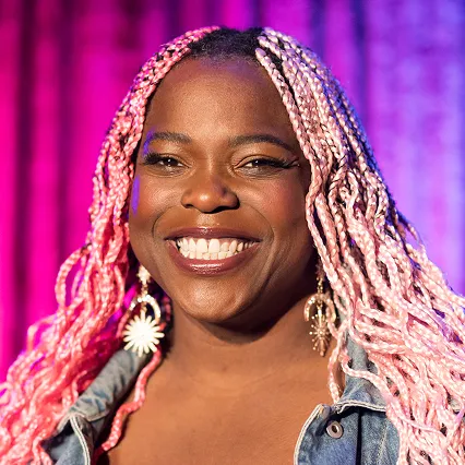 Tamara Shevon, stand-up comedian, smiles in a portrait against a purple and blue stage backdrop.