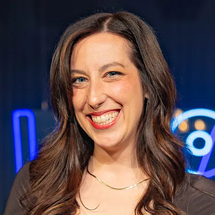 Alexa Stanton, stand-up comedian, smiles in a portrait against a dark stage backdrop with blue neon lights.