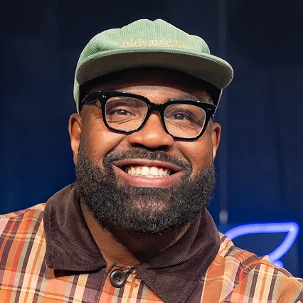 Dave Helem, stand-up comedian, smiles in a portrait wearing glasses and a green cap against a dark stage backdrop with blue neon lights.