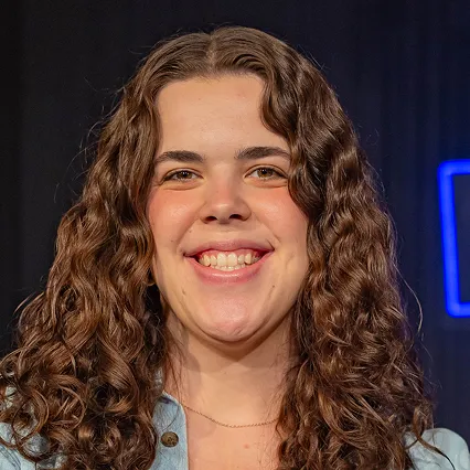 HannahBeck Hannah Beck, stand-up comedian, smiles in a portrait against a dark stage backdrop with blue neon lights.