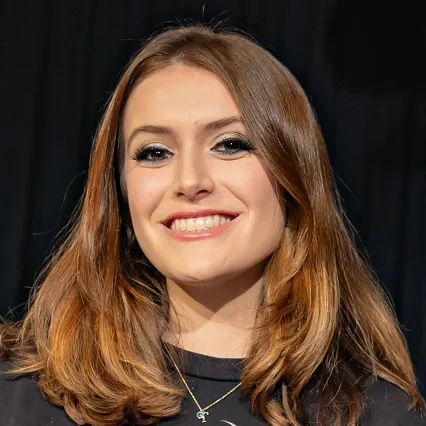 Hannah Belmont, stand-up comedian, smiles in a portrait against a dark stage backdrop.