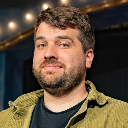 HunterHirsh Hunter Hirsh, stand-up comedian, poses in a portrait against a dark stage backdrop with string lights overhead.