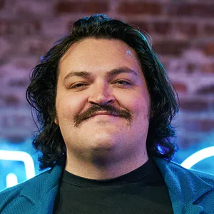Smiling portrait of comedian Colin Armstrong in front of a blue neon LMAOF sign.