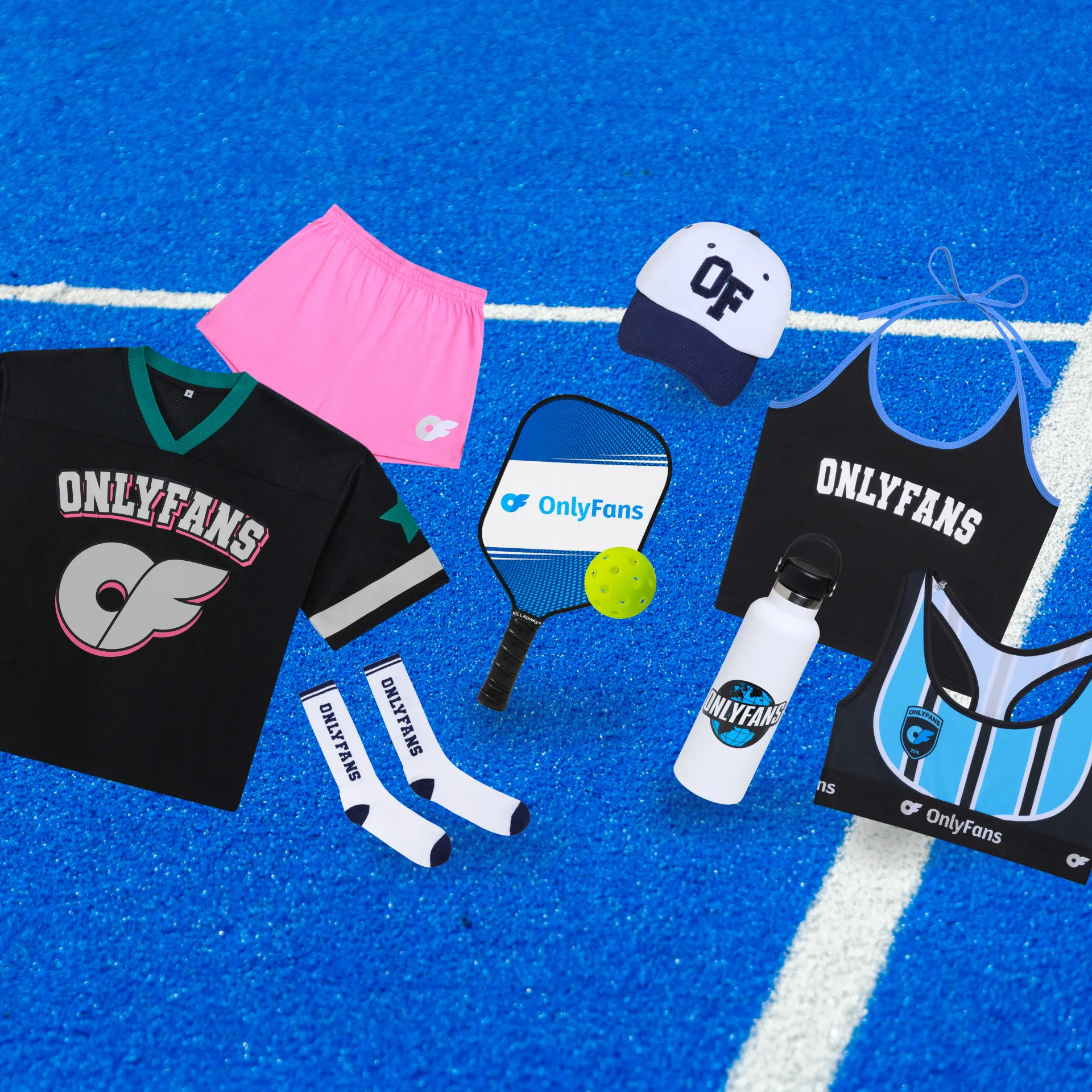 Img2_AllScreenSizes_1920x1920 V 5 OnlyFans pickleball paddle displayed alongside pieces from The Stadium Kit collection, including a jersey, shorts, cap, socks, water bottle, sports bra, and tote bag on a blue pickleball court.