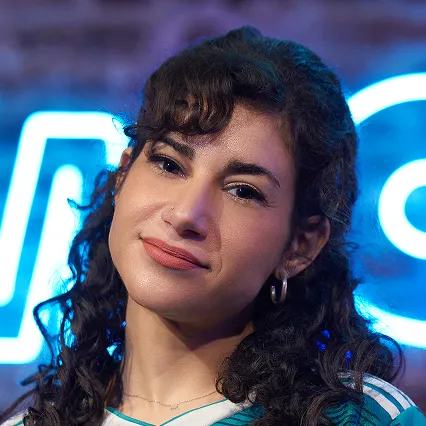 Portrait of comedian Naomi Karavani in a black high-neck top in front of a blue neon LMAOF sign.