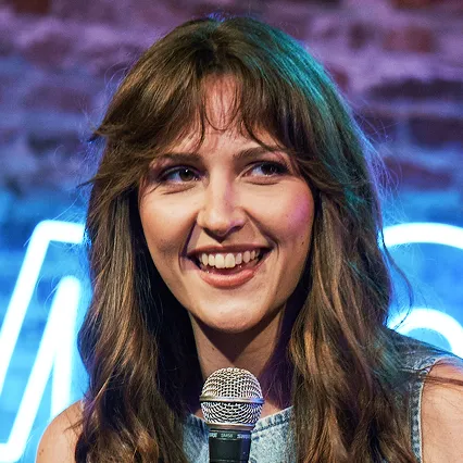Smiling portrait of comedian Stephanie Robertson holding a microphone in front of a blue neon LMAOF sign.