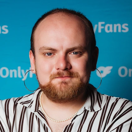 AndrewWhite Comedian Andrew White poses in a striped shirt and hoop earrings in front of an OnlyFans backdrop.