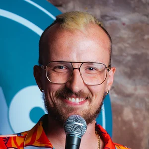 Comedian Andy Field smiles behind a microphone while wearing glasses and a bright patterned shirt in front of an LMAOF backdrop.