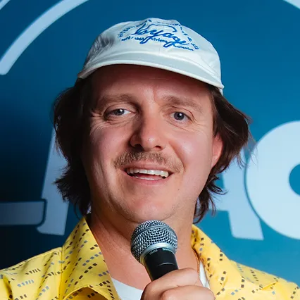British comedian Joey Page smiles while holding a microphone and wearing a white cap and yellow shirt in front of an LMAOF backdrop.
