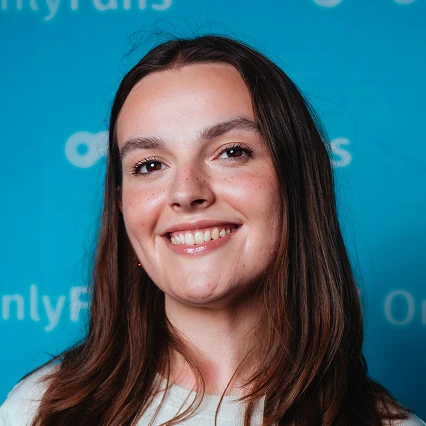 MillieMalone British comedian Millie Malone smiles while posing in front of an OnlyFans backdrop.